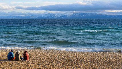 Incline Beach - Young tourists enjoying a sunny Spring evening at Incline Beach, as storm clouds moving over snow peaks into Lake Tahoe. California-Nevada, USA. Focus is on sandy beach in foreground.
