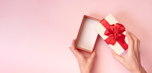Hands holding open gift box with red bow on pink background. Top view