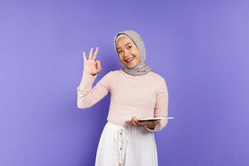 An excited beautiful Asian woman in a pink sweater and hijab holding a tablet and making an OK sign, received good news isolated over a purple background