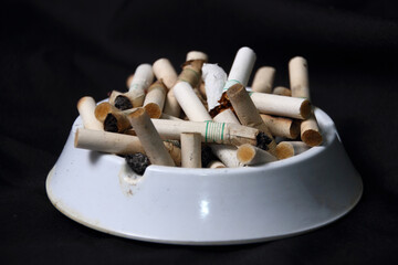 Pile of cigarette butts in white ashtray on dark background, smoking addiction concept
