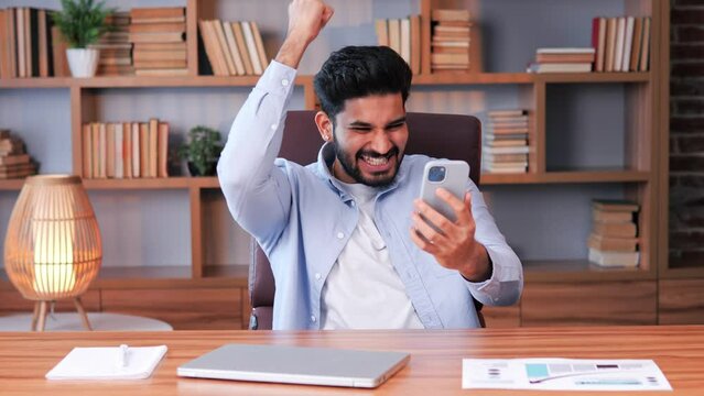 Successful Hindi businessman in office looking at mobile phone with an expression of triumph or victory, perhaps related to financial stocks, sports betting or a game he is playing. Success happiness.