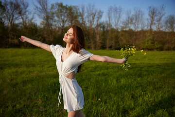 Naklejka na ściany i meble a girl in a light summer dress stands in a field with a bouquet of flowers raising her hands up