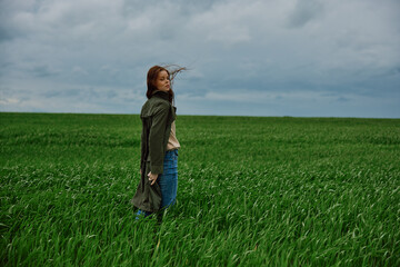 a woman in a coat walks through a green field in rainy weather. Strong wind, harmony with nature