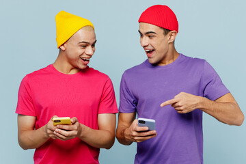 Young cheerful fun happy couple two friends men wearing casual clothes together hold in hand use point index finger on mobile cell phone show memes isolated on pastel plain light blue cyan background. - obrazy, fototapety, plakaty