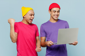 Young fun couple two friends IT men wear casual clothes together hold use work on laptop pc computer do winner gesture clench fist isolated on pastel plain light blue cyan background studio portrait.