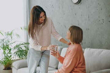 Two smiling adult women mature mom young kid wear casual clothes give glass of water to drink sit on gray sofa couch stay at home flat rest relax spend free spare time in living room. Family concept.