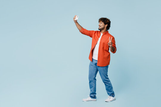 Full body fun young Indian man wear orange red shirt white t-shirt doing selfie shot on mobile cell phone post photo on social network show v-sign isolated on plain pastel light blue cyan background.