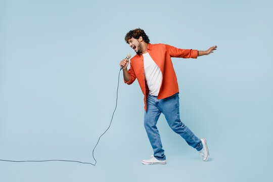 Full body side profile view happy young Indian man wear orange red shirt white t-shirt sing song in microphone isolated on plain pastel light blue cyan background studio portrait. Lifestyle concept.