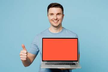 Young smiling happy IT man wear casual t-shirt hold use work on laptop pc computer with blank screen workspace area show thumb up isolated on plain pastel light blue cyan background studio portrait.