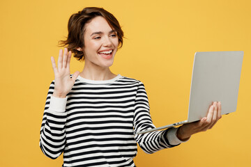 Young smiling happy fun IT woman wearing casual striped shirt hold use work on laptop pc computer waving hand get video call talk isolated on plain yellow color background studio. Lifestyle concept.