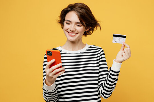 Young happy woman wear casual striped shirt using mobile cell phone hold credit bank card doing online shopping order delivery booking tour isolated on plain yellow color background studio portrait.