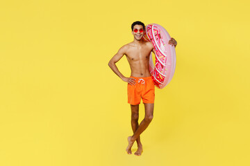 Full body happy young sexy man wear orange shorts swimsuit relax near hotel pool hold on shoulder pink donut rubber ring isolated on plain yellow background. Summer vacation sea rest sun tan concept.