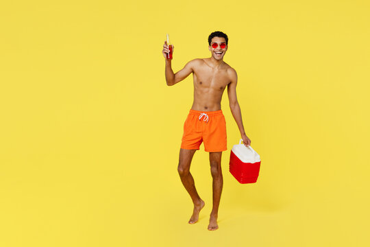 Full body fun young man wear shorts swimsuit glasses hold in hands beer bootle beach refrigerator relax near hotel pool isolated on plain yellow background. Summer vacation sea rest sun tan concept.