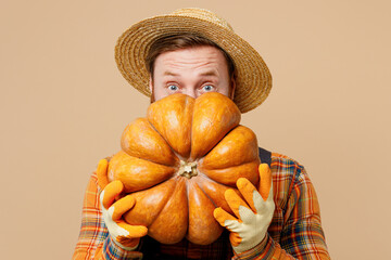 Happy young bearded man wearing straw hat overalls work in garden hold cover face with pumpkin look...