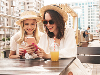 Two young beautiful smiling hipster female in trendy summer white dress and hats. Sexy carefree women drinking fresh vegetable cocktail smoothie drink in plastic cup with straw. Using phone apps