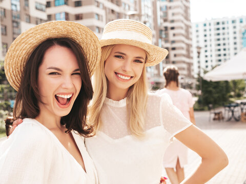 Two young beautiful smiling hipster female in trendy summer white dress clothes and straw hats. Sexy carefree women walking in street. Positive models having fun, hugging and laughing