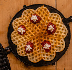 An antique waffle iron with a freshly baked waffle in it decorated with raspberry jam and creamth...