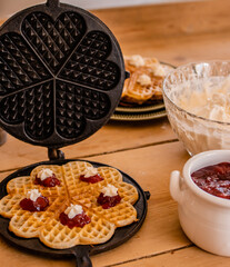 An antique waffle iron with a freshly baked waffle in it decorated with raspberry jam and cream next to it there is a bowl with jam and one with cream