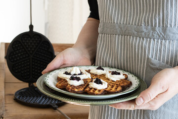 A lady in a blue striped apron holds a plate with a waffle garnished with strawberry jam and cream.