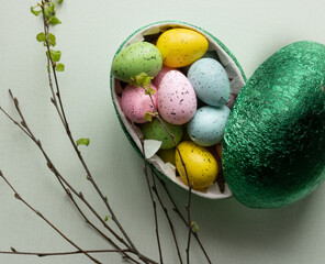  A green Easter egg filled with green, blue, yellow and pink Easter caramels. Nearby there is a birch twig on a light background