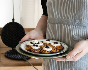  A lady in a blue striped apron holds a plate with a waffle garnished with strawberry jam and cream.