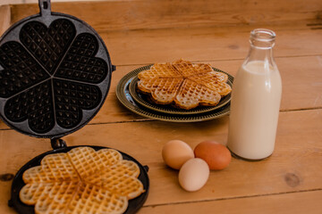 An antique waffle iron with a freshly baked waffle next to it are country eggs, milk bottle and a dish with freshly baked waffles