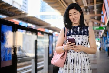 An attractive young confident Asian business woman is using her cellphone while waiting for her train at a station to go to work in the morning.