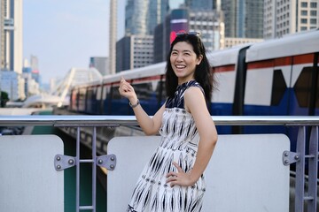 An Asian woman is standing on a platform at a sky train station, feeling happy and smiling with the incoming train arriving in the background.