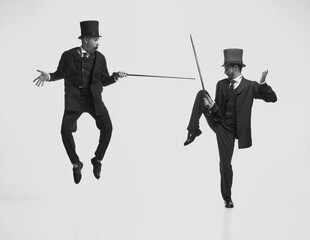 Black and white portrait of two gentlemen wearing classic suits jumping and fighting with canes...
