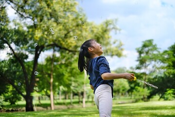 A cute young Asian girl is playing with bubbles using a bubble wands in a park on a bright sunny...