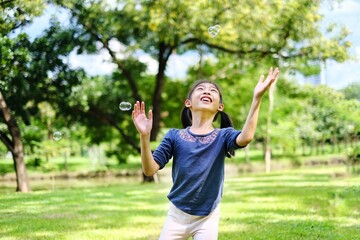 A cute young Asian girl is looking up and playing with bubbles in the park with beautiful trees and green grass.