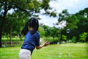 The back side of a cute young Asian girl running, spinning and trying to catch bubbles in a park with beautiful trees and green grass in the background.