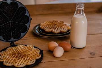 An antique waffle iron with a freshly baked waffle next to it are country eggs, milk bottle and a dish with freshly baked waffles