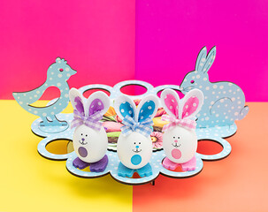 Easter concept. Festive stand for Easter eggs. eggs with painted muzzles and ears made of fabric. Rabbit and chicken made of wood on a bright multi-colored background.