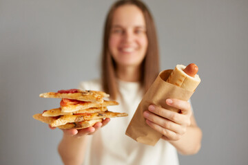 Portrait of brown haired attractive woman holding offering hotdog and pizza, inviting you to eat junk food standing isolated on gray background wearing white t-shirt.
