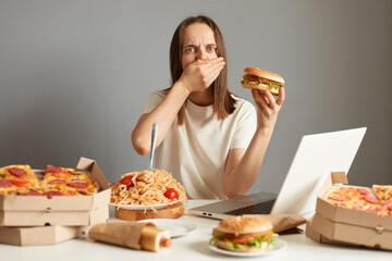Portrait of sick overeating woman sitting at table in front of notebook isolated over gray background, holding sandwich, feels bad and nausea, covering mouth with palm.
