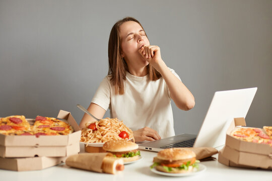 Image of sleepy tired woman working or watching movie on laptop during long hours isolated over gray background, being surrounded with pizza, pasta, hotdogs, hamburgers.