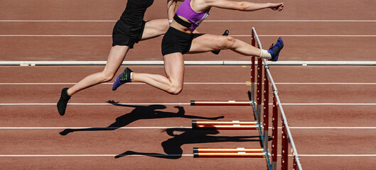 two female athletes running hurdles in athletics competition, hurdling on stadium track, summer...