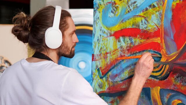 Creative individual is an artist who is creating a masterpiece of art in the studio. The man is painting on canvas with oil paints, while listening to his favorite music through white headphones.