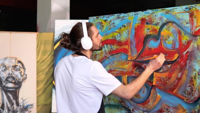 Imaginative artist with long hair using skill innovation to craft work of art. Using red, blue, yellow paints, artist is producing a magnificent painting on a sizable canvas that is sure to impress.