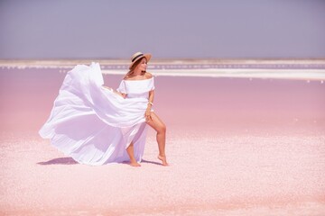 Fototapeta na wymiar Woman in pink salt lake. She walks in a white long dress and hat along the salty white shore of the lake. Wanderlust photo for memory