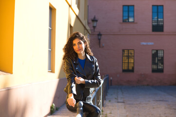 Fototapeta na wymiar Young and beautiful Hispanic brunette woman with curly hair and black leather clothes is sitting on a railing in the street. The woman is happy and smiling.
