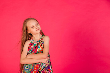 Dreaming little girl in colorful t-shirt posing arms crossed at pink background, looking up away. Young dreamy lady with long hair expressing emotion. Childhood emotional concept. Copy ad text space