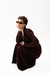 young model in brown pantsuit and sunglasses posing with hand near face on white background.