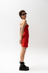 Fototapeta premium full length of slender tattooed woman in sunglasses and red strapless dress with black boots on grey background.