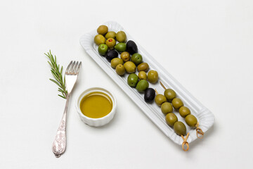 Green olives on skewers. Olives on white plate. Olive oil in bowl.