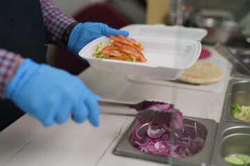 Cook preparing healthy lunch meal for take away in a Greek restaurant. Kitchen worker preparing traditional kalamaki dish with fresh vegetables