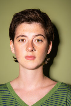 Portrait Of Brunette Woman With Freckles And Nose Piercing Looking At Camera On Green Background.