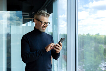 Mature adult man inside office standing near window, businessman typing message on phone and browsing internet pages, gray-haired boss smiling reading news online.