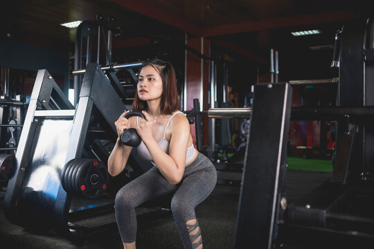 A young asian woman does a set of goblet squats with a kettlebell. Holding the weight in front of her chest. Training at a modern gym.